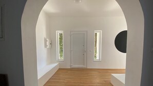 A spacious room with a wooden floor, white walls, and a white door. There are two windows with greenery visible outside.