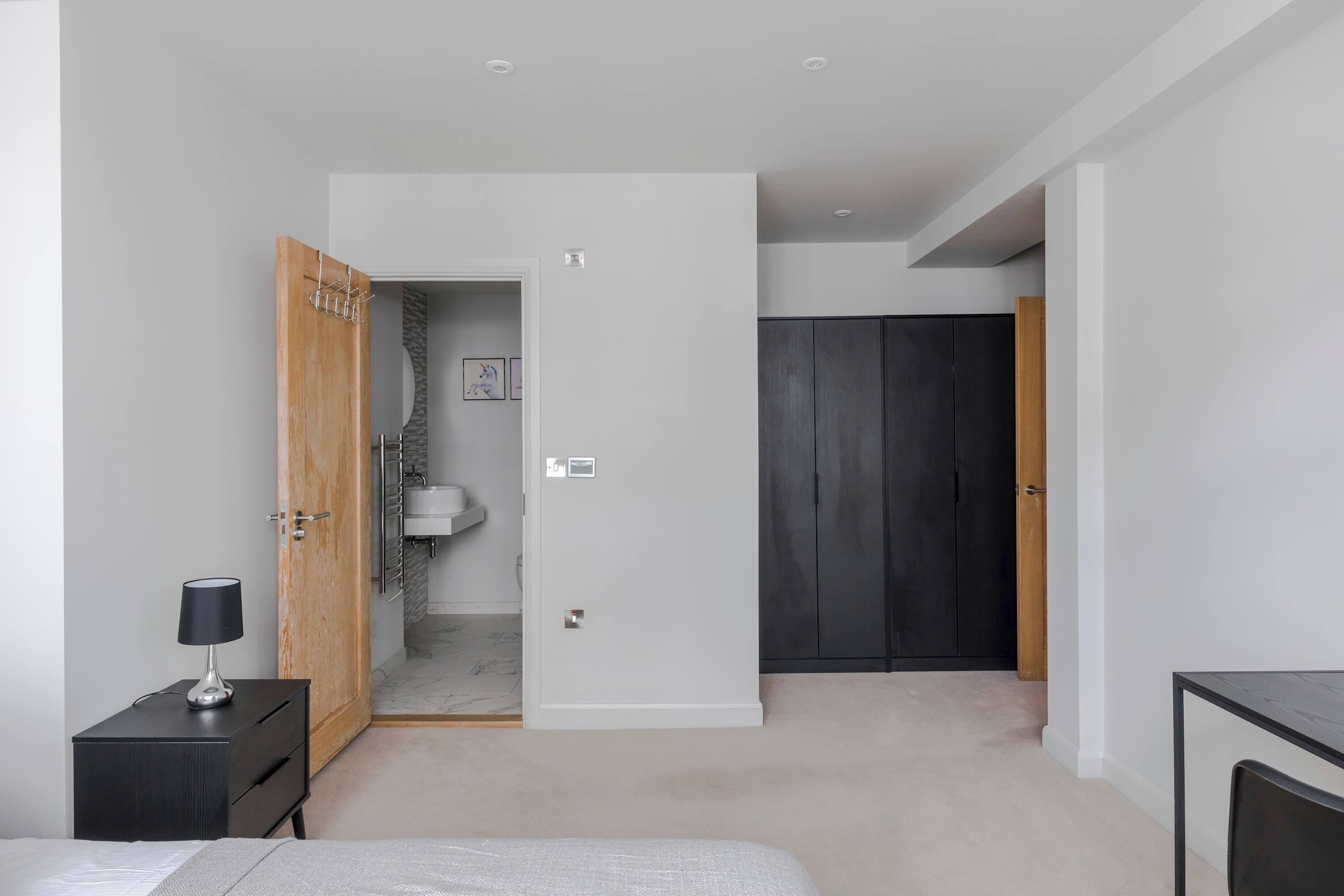A bedroom with a bed, bedside table, lamp, wardrobe, and a bathroom visible through an open door.