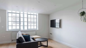 A modern living room with a sofa, a small round table, a TV mounted on the wall, and a large window providing natural light. - Clerkenwell 2-Bed · 2 Baths · Sleeps 6 (London)