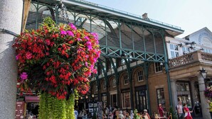 Un edificio histórico con estructura metálica verde, un canasto de flores y un letrero para Colbert Garden Market.