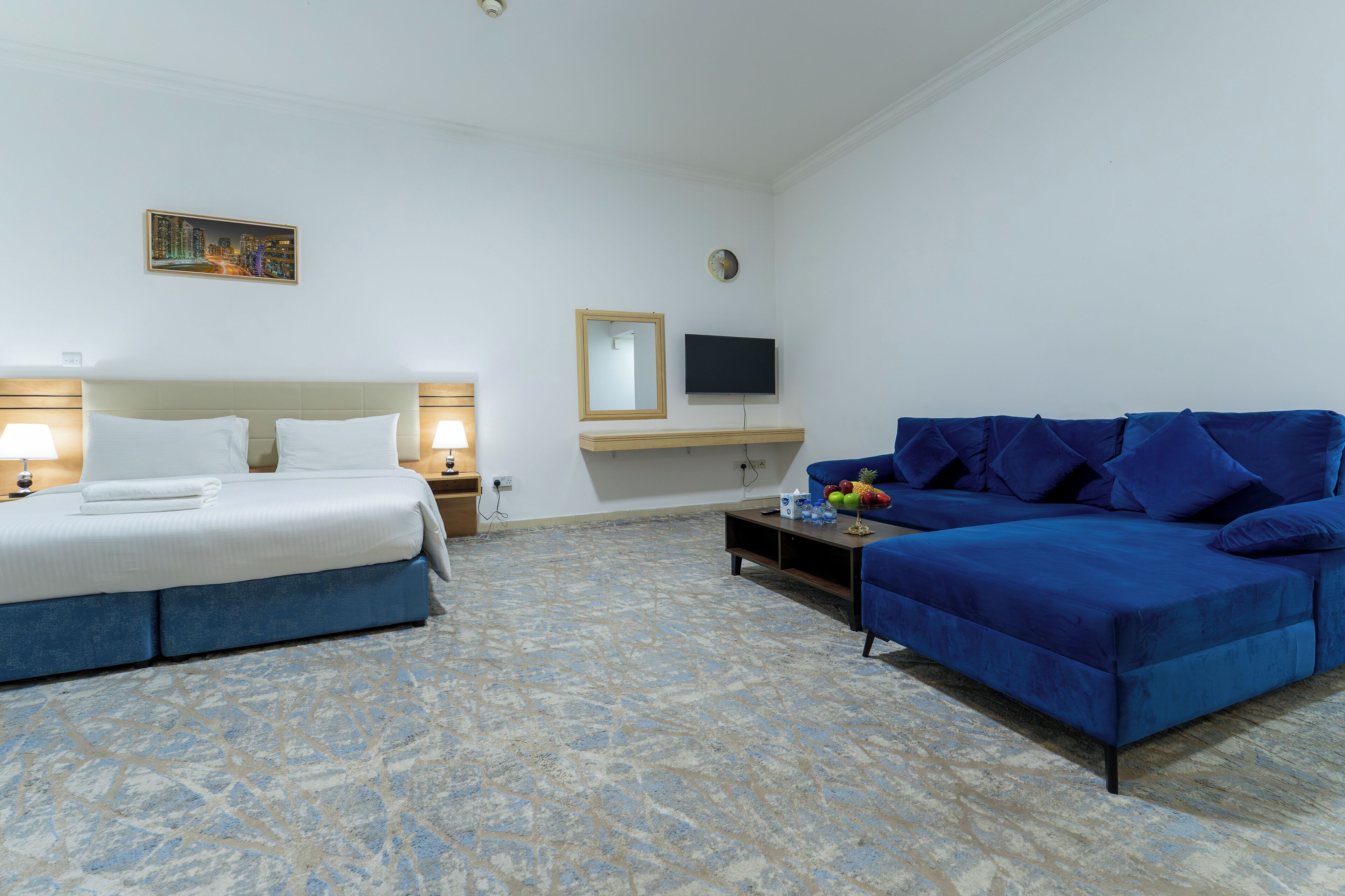 A hotel room with a bed, a blue sofa, a TV, a mirror, and a clock on the wall.