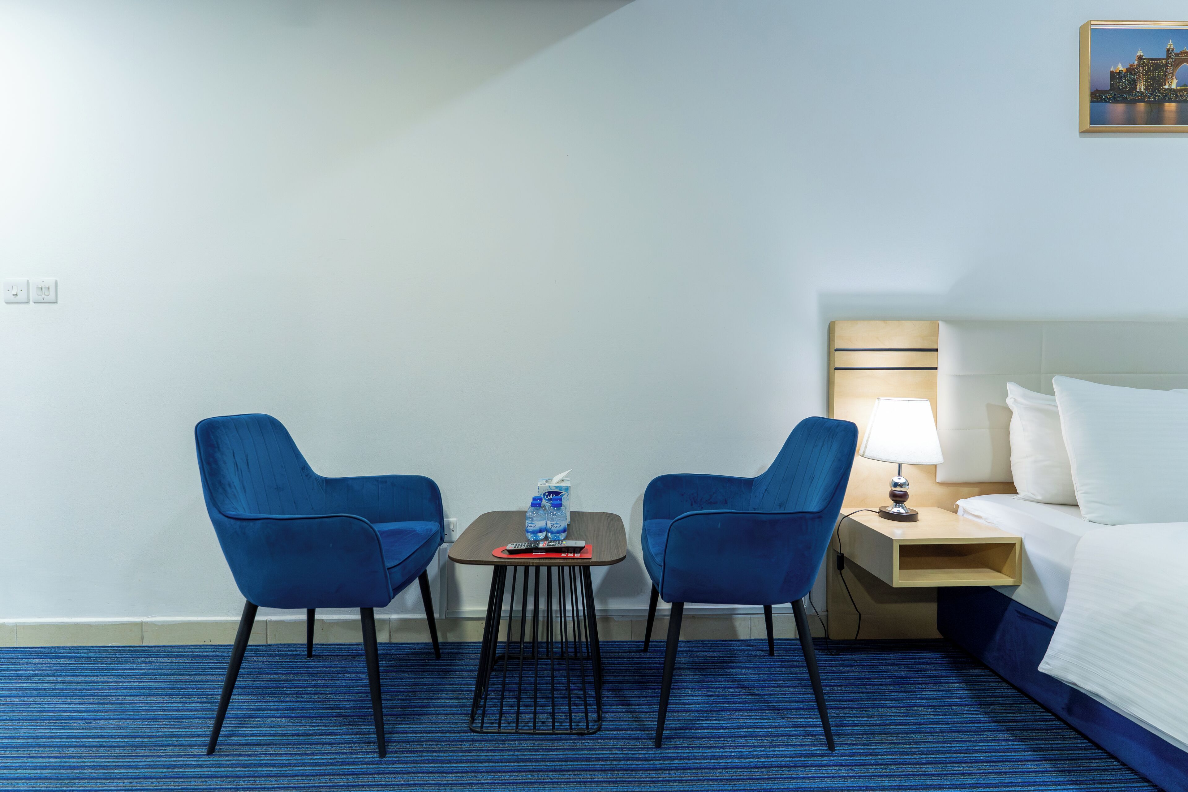 Ein Hotelzimmer mit einem Bett, zwei blauen Stühlen, einem kleinen Tisch mit Fernbedienung, einer Wasserflasche und einer Lampe.