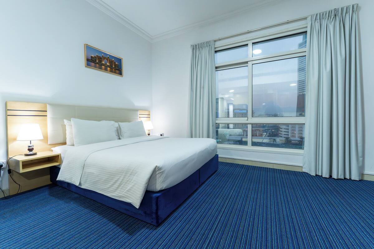 Hotelkamer met een groot bed, nachtkastje, lamp, raam met gordijnen en uitzicht op de stad.