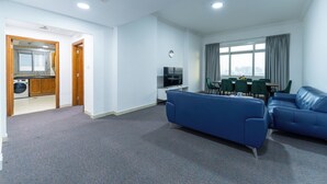Uma sala de estar moderna com um sofá azul, uma área de jantar com cadeiras verdes e uma cozinha visível através de uma porta aberta.