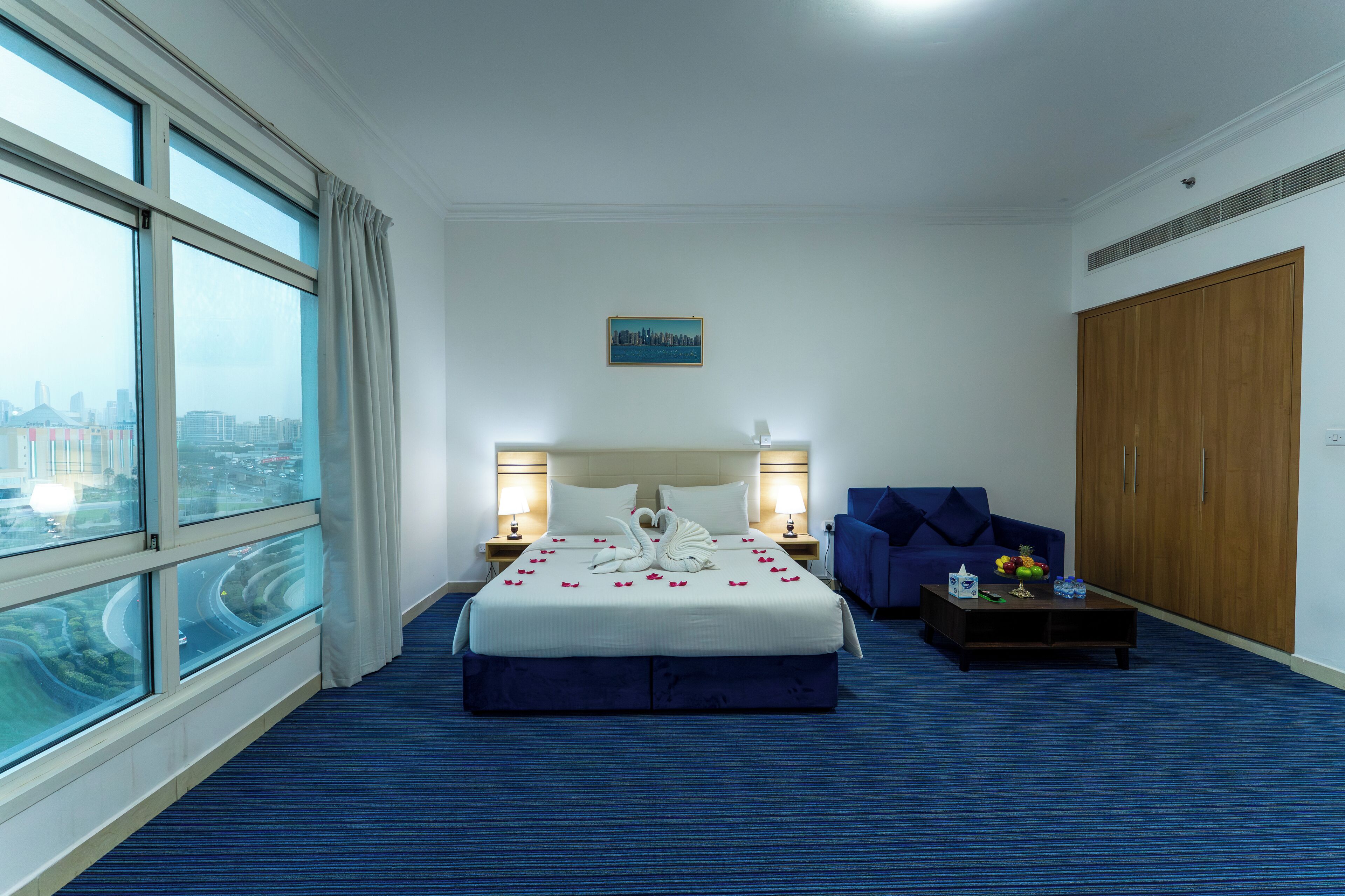 A hotel room with a large bed, two bedside lamps, a view of a cityscape through large windows, a blue sofa, and a wooden wardrobe.