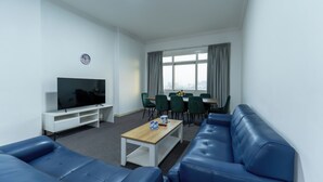 Uma sala moderna com televisão de tela plana, sofás de couro azul, uma mesa de centro de madeira e um espaço de jantar com cadeiras verdes.