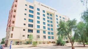 Um hotel com vários andares, fachada bege, inúmeras janelas e área ajardinada com árvores e arbustos.