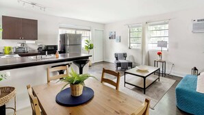 A modern kitchen with a central island, a dining area with a wooden table and chairs, a sofa, and a small side table with a lamp. - Brickell Pearl Cozy House/Beach 10 min/Location (Miami)