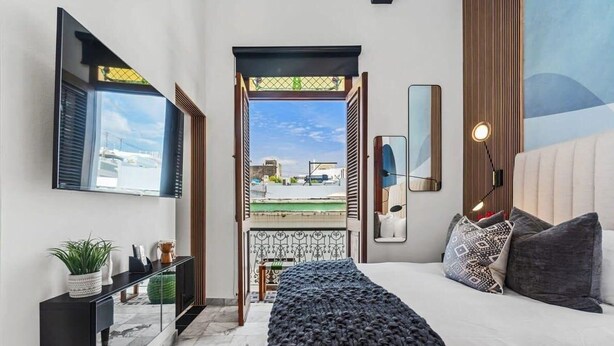 A modern bedroom with a large bed, a nightstand, a mirror, and a view of the city through an open door. - Villa Mosaico | Stylish & Modern Boutique 1 BR (San Juan)