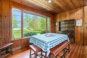 A wooden-paneled room with a table set for two, a bench, and a view of a green landscape through large windows.