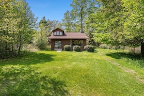 A small, single-story house with a sloped roof, surrounded by a well-maintained lawn and dense trees.