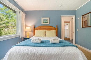 A bedroom with a wooden bed, a window with curtains, a bedside lamp, and a framed picture on the wall.