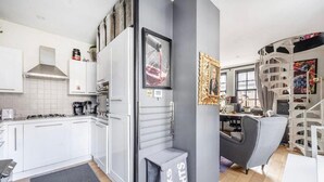 Ett modernt kök med vita skåp, en inbyggd ugn och en kaffebryggare. Det ligger en grå matta på golvet och en papperskorg med texten ”DARKS LIGHTS”.