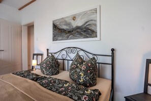 A bedroom with a bed, floral-patterned pillows, a wall-mounted painting, and a bedside lamp.