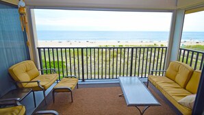 Un balcon avec vue sur la plage, des chaises jaunes, une table et un luminaire suspendu.