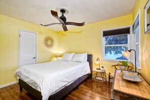 A bedroom with a bed, a ceiling fan, a window with blinds, a bedside table with a lamp, and a small plant. - Walk 2 Beach, EV Charger, Outdoor Dining, 2 Units (Carlsbad)