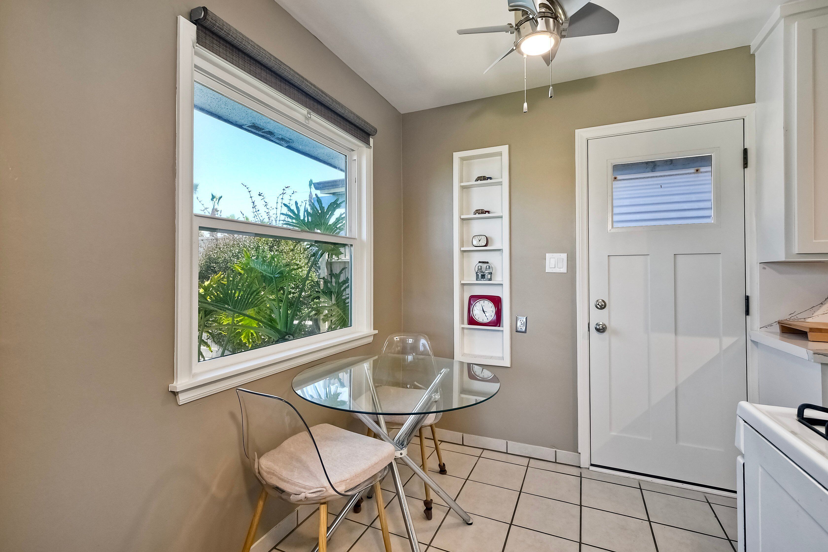Une petite cuisine avec une table et des chaises en verre, une porte blanche et une fenêtre avec des stores.