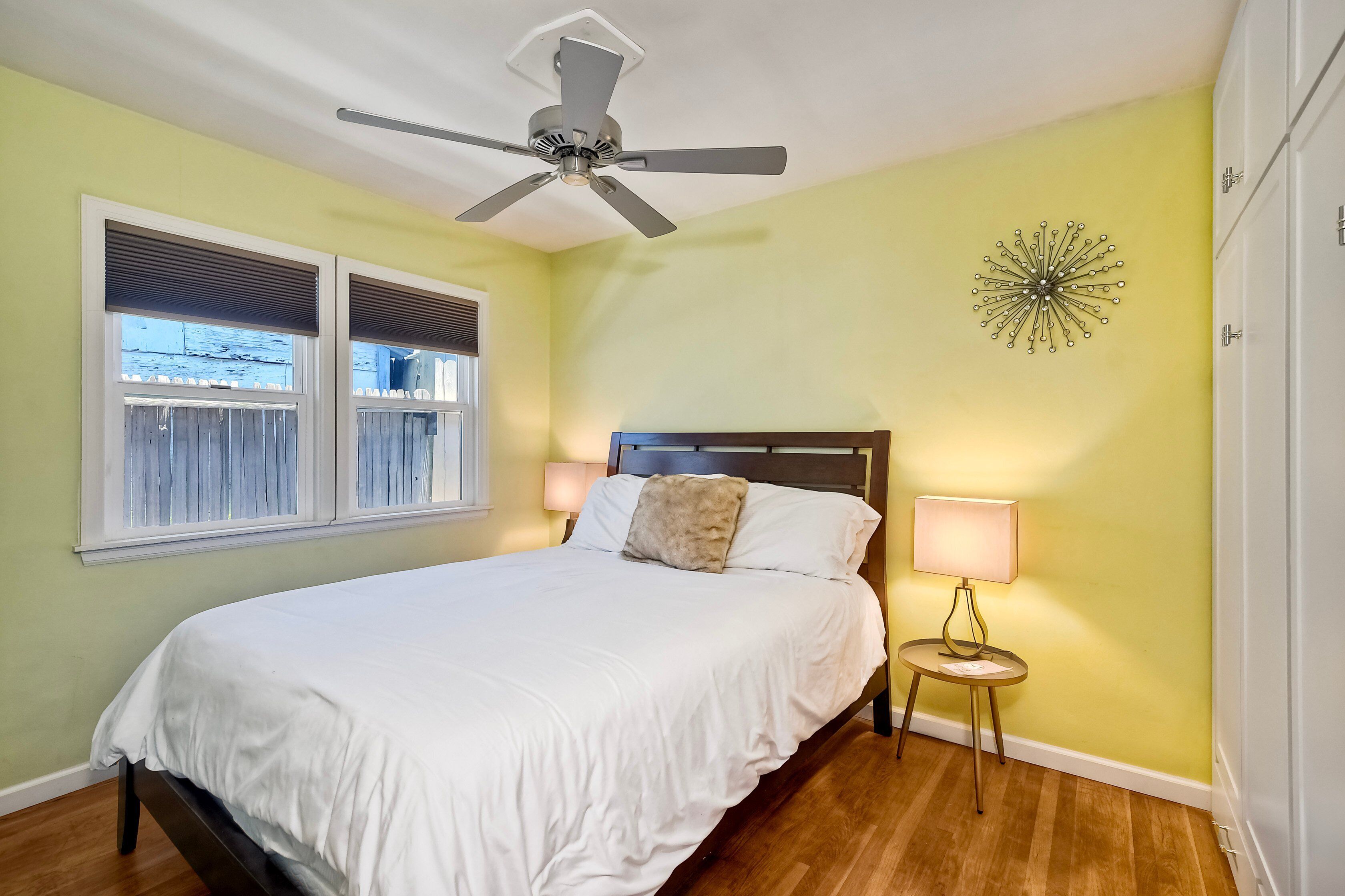 A bedroom with a bed, a ceiling fan, a window with blinds, a wall-mounted lamp, and a decorative wall piece.