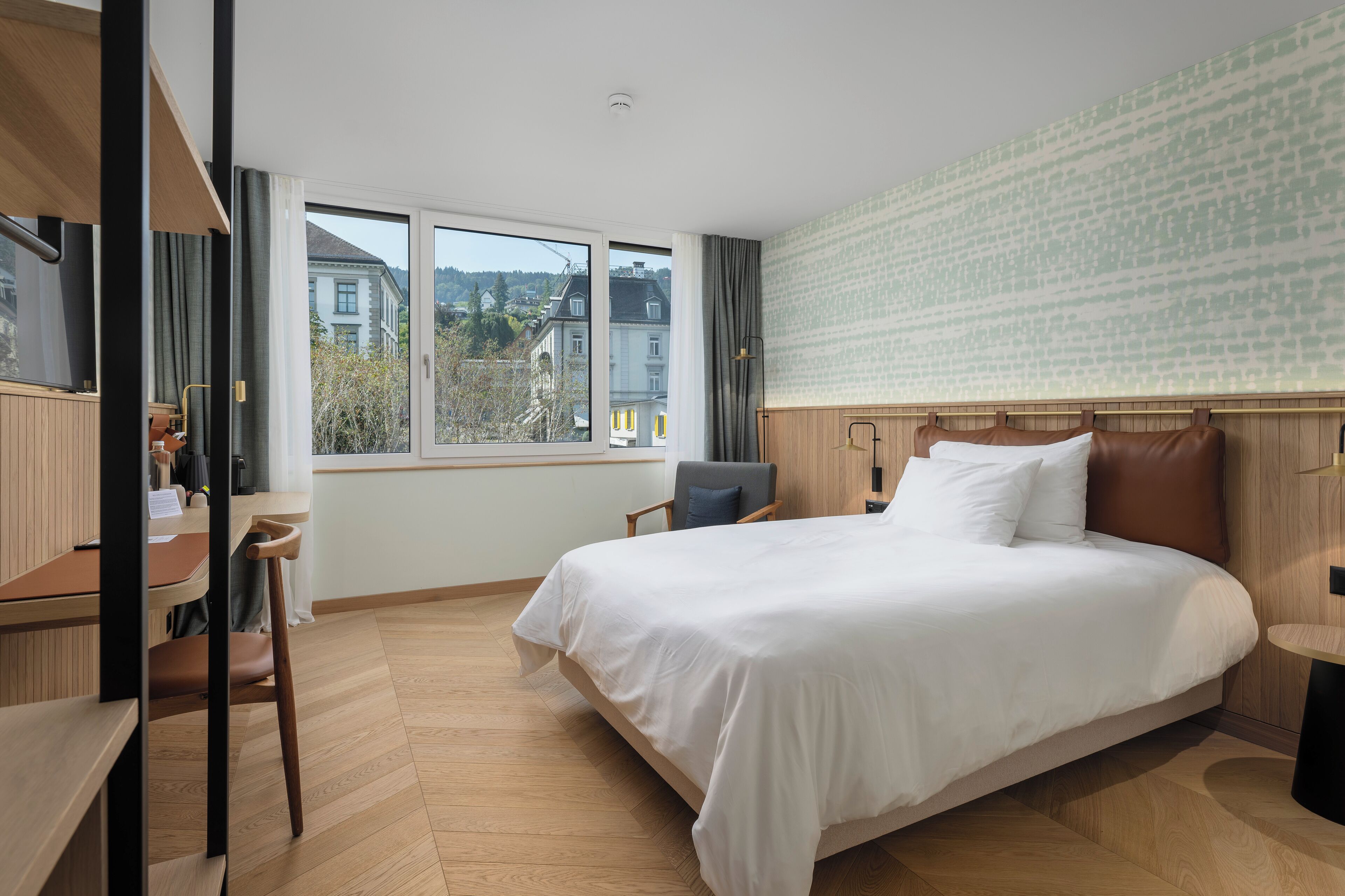 A modern hotel room with a large bed, a desk, and a view of buildings through the window.