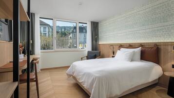 A modern hotel room with a large bed, a desk, and a view of buildings through the window.