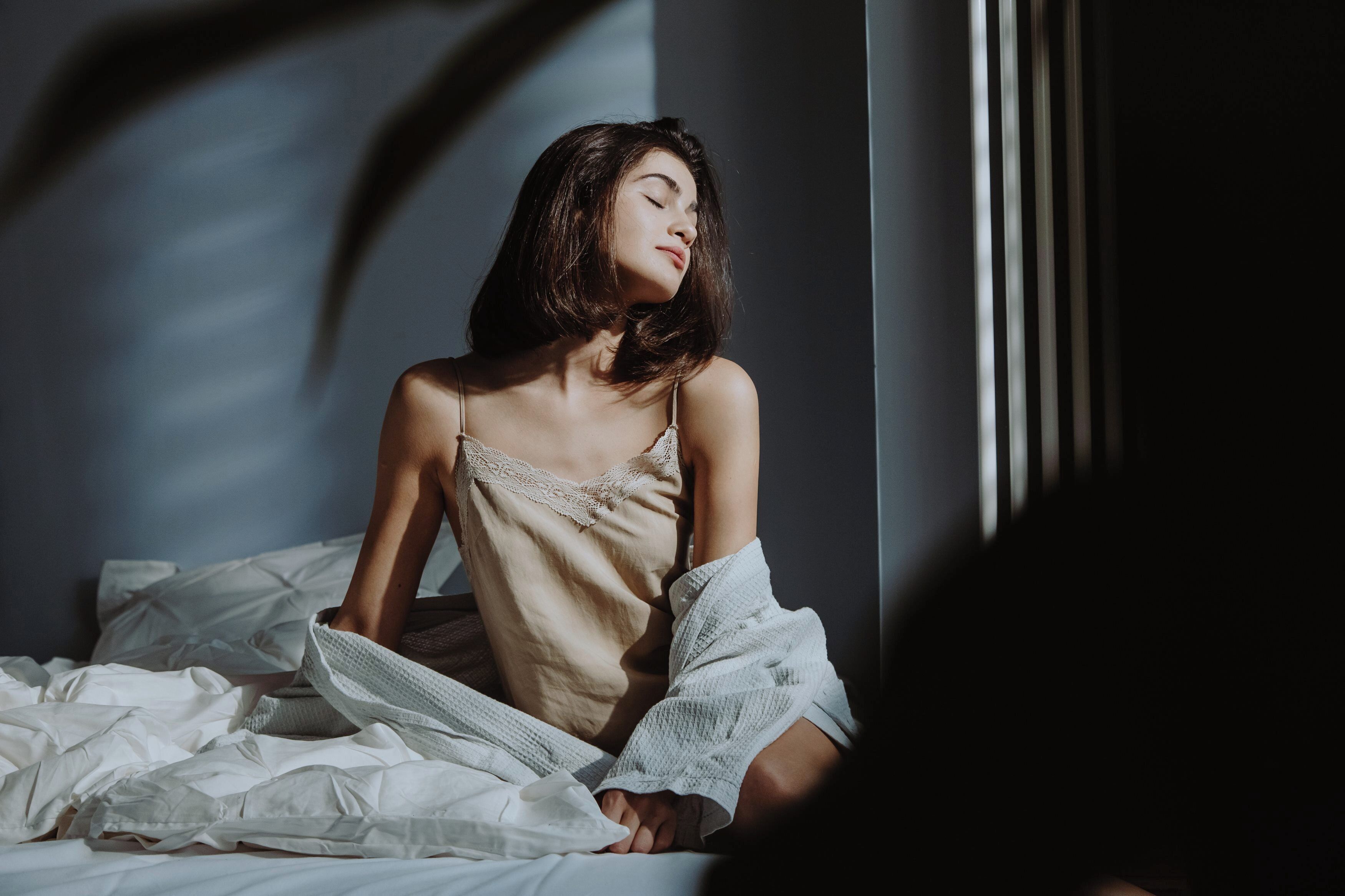 Una persona sentada en una cama con sábanas blancas, vestida con una blusa color beige y una bata blanca.