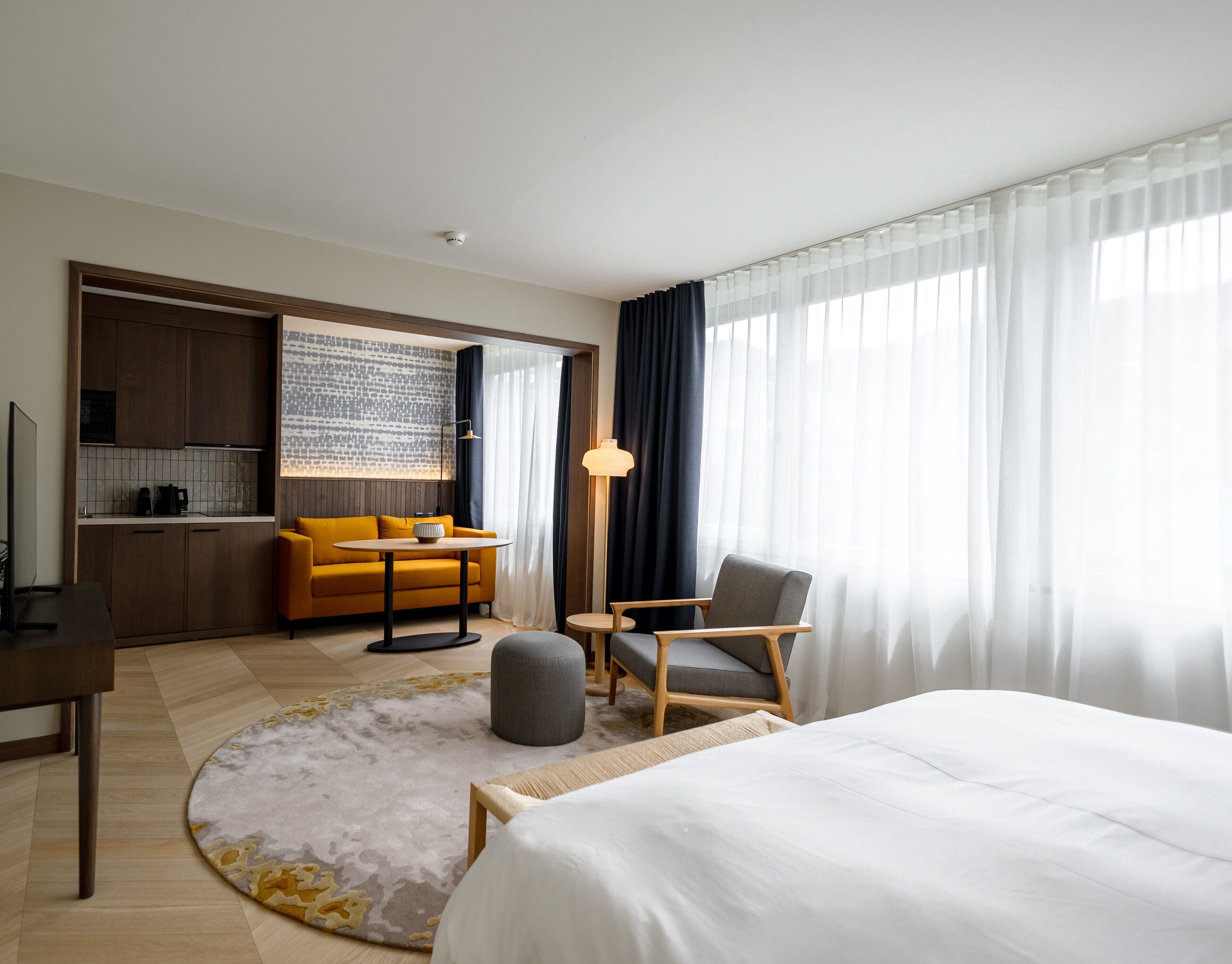 A modern hotel room with a bed, a sofa, a chair, a small table, a lamp, and a kitchen area with a coffee machine.