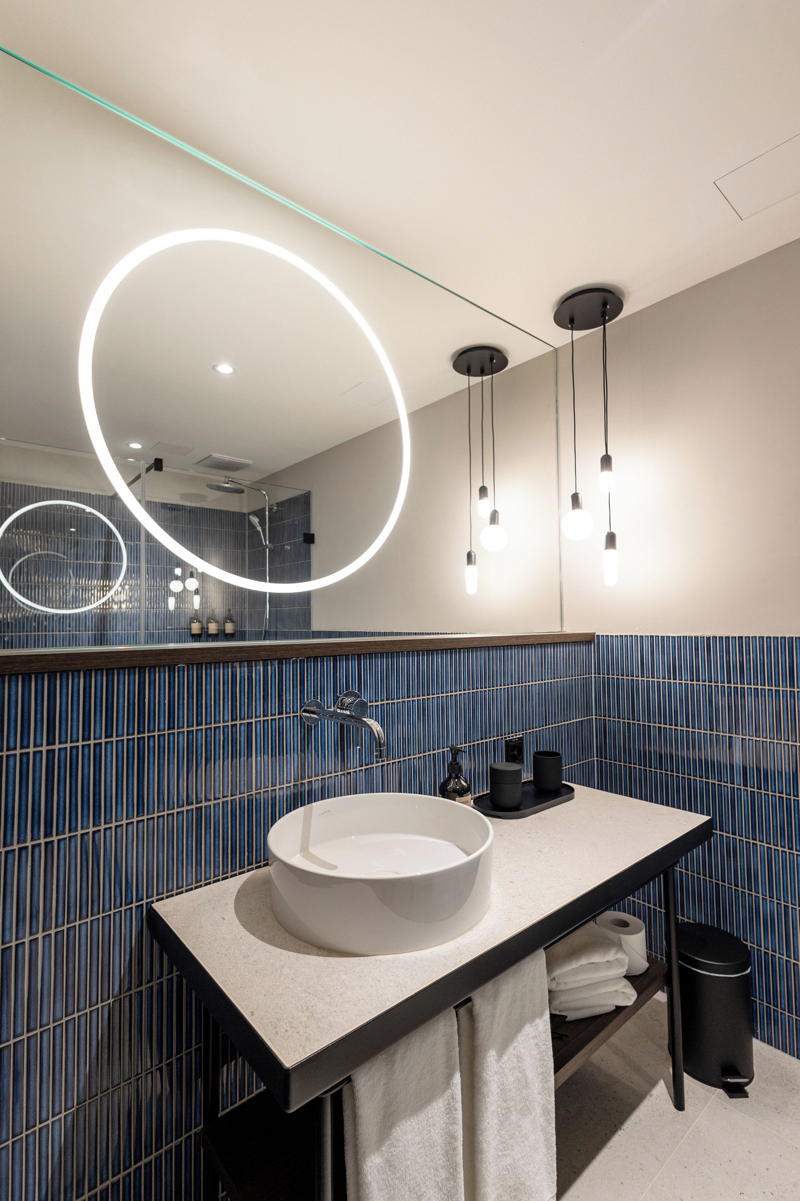 Une salle de bain moderne avec un grand miroir rond, un lavabo blanc et des murs recouverts de carreaux bleus.