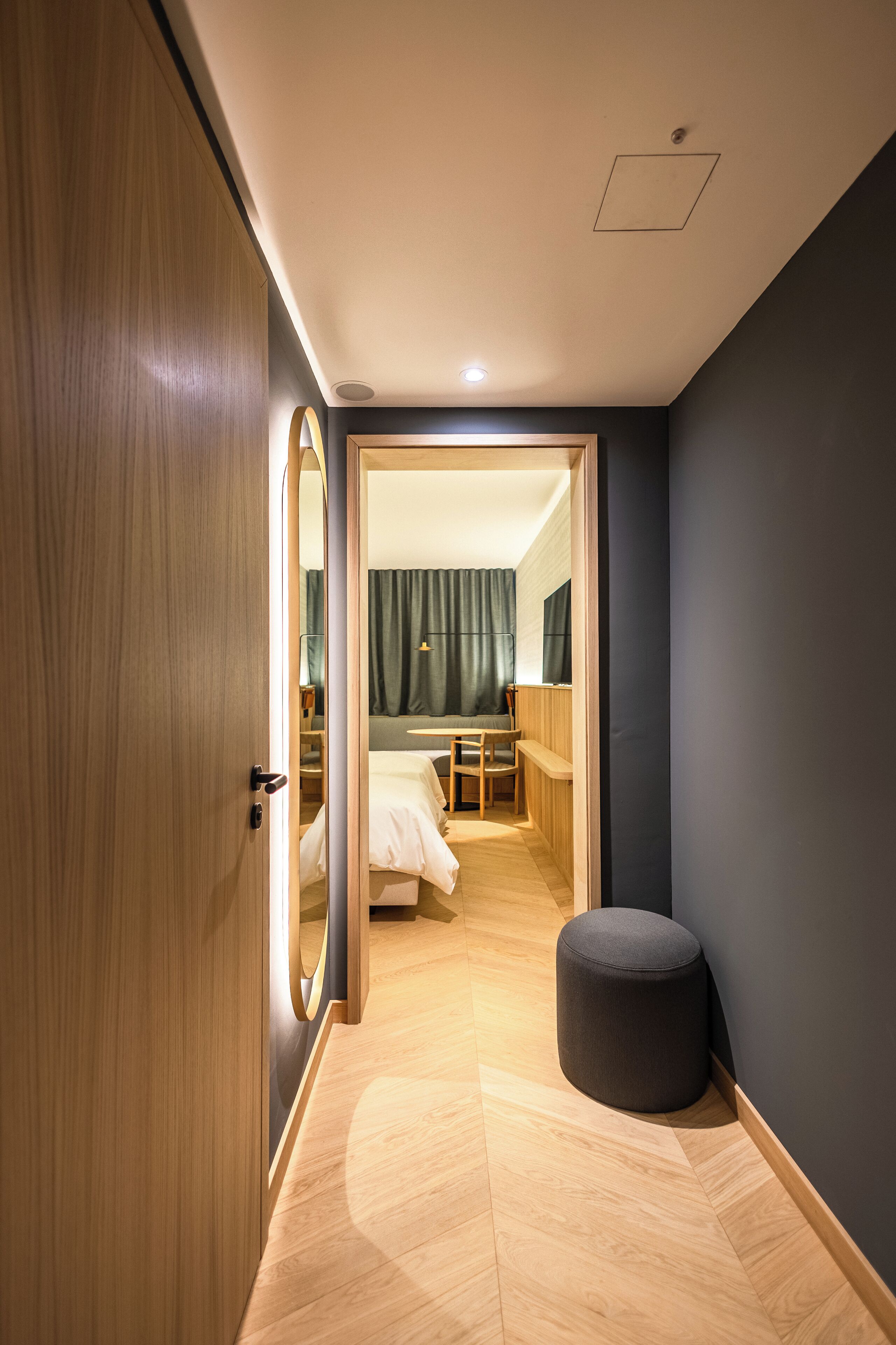 Una habitación de hotel moderna con un armario de madera, un otoman redond, una cama, un escritorio y un espejo.