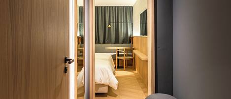 Ein modernes Hotelzimmer mit Holzschrank, rundem Ottomanen, einem Bett, einem Schreibtisch und einem Spiegel.
