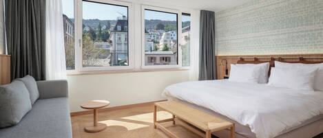 Habitación de hotel moderna con una cama grande, un sofá gris, una mesita redonda y un banco. La habitación tiene ventanas amplias con vistas a edificios y árboles.