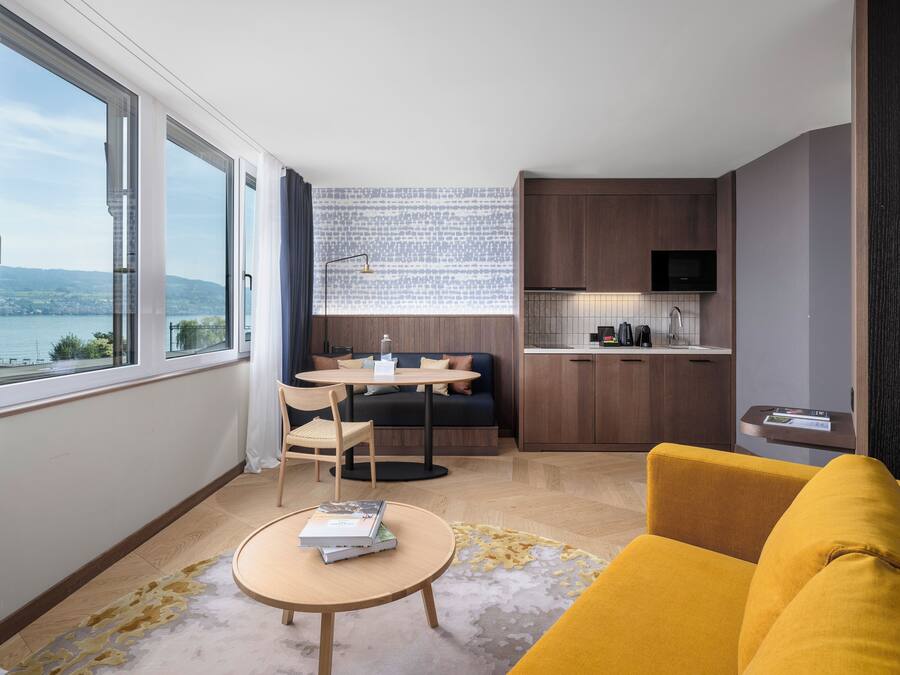 Una sala de estar moderna con un sofá amarillo, una mesa de centro redonda de madera y vistas a un paisaje urbano a través de amplios ventanales.