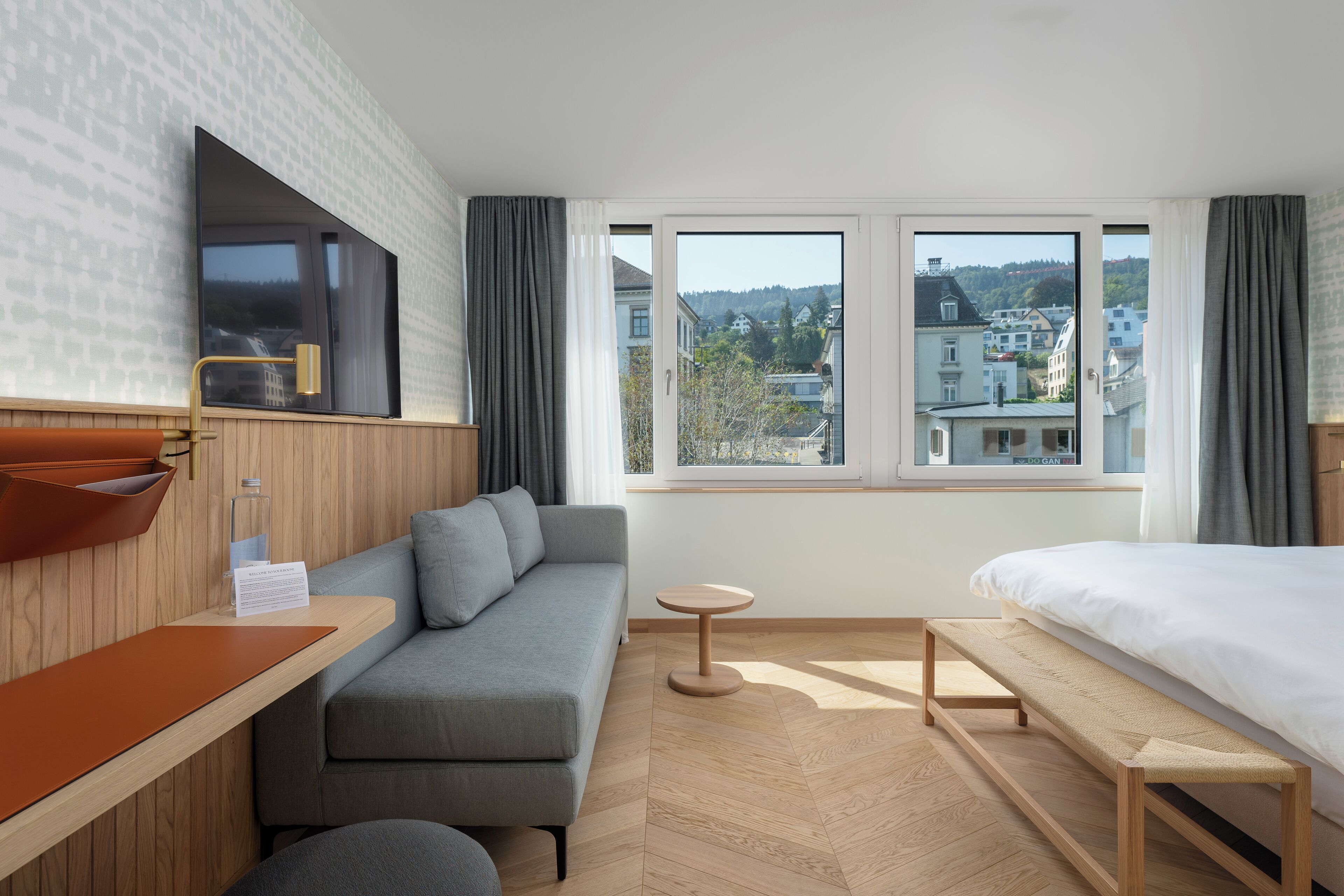 Une chambre d’hôtel moderne, dotée d’un canapé, d’un lit, d’une petite table et offrant une vue sur la ville grâce à de grandes fenêtres.