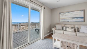 A room with a large sliding glass door leading to a wooden deck with a view of the beach. There is a beige sofa with patterned cushions and a painting above it.