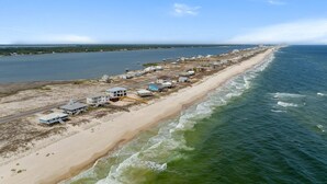 An aerial view of a coastal area with a long stretch of beach and ocean. There are several buildings along the shore, some with visible structures like staircases and driveways.