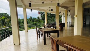 An indoor dining area with tiled flooring, wooden tables and chairs, hanging pendant lights, and a view of lush greenery through large windows. - Red Letter Hotel (Kandy)