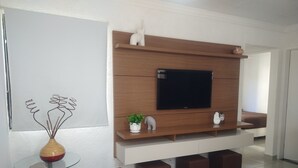 Un salon moderne avec un téléviseur à écran plat fixé sur un mur recouvert de panneaux en bois, une table basse en verre, un canapé beige et un vase décoratif contenant une plante.