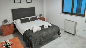 Una cama bien hecha con cabecera gris, dos cuadros enmarcados en la pared, una mesita de noche con lámpara y una ventana con vistas al cielo.