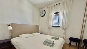 A neatly made bed with pillows and folded towels, a bedside lamp, a clock on the wall, and a window with curtains. - Beautiful 2BR In Ramat Gan (Ramat Gan)