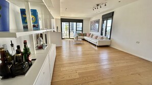 A spacious living room with a white sofa, wooden flooring, and a large window offering a view of the city. - Beautiful 2BR In Ramat Gan (Ramat Gan)