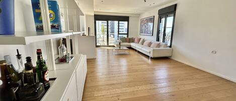 A spacious living room with a white sofa, wooden flooring, and a large window offering a view of the city.