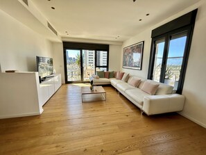 Un salon moderne avec un canapé blanc, une table basse et un téléviseur posé sur un meuble blanc. De grandes fenêtres offrent une vue sur la ville.