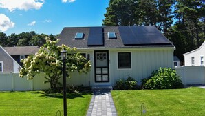 Une maison de plain-pied avec une allée pavée, des panneaux solaires sur le toit et un jardin devant la maison, avec un arbre et un petit arbuste.