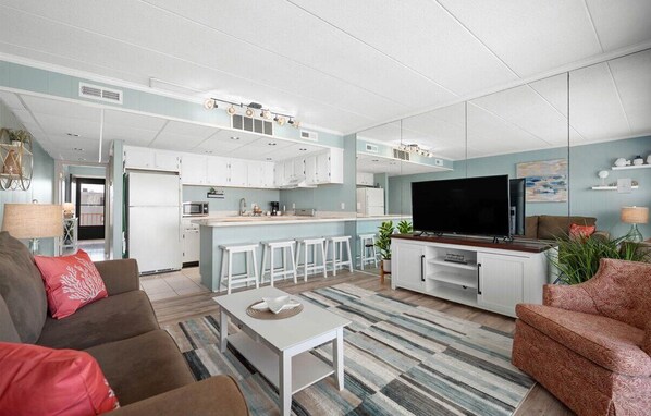 A modern living room with a sofa, a coffee table, and a TV. There is a kitchen area with white cabinets and a bar counter. - Updated Mid-Town Ocean Block Unit - Ready for Summer Fun! (Ocean City)
