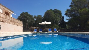 Une piscine d’un bleu éclatant, bordée de carreaux, entourée de chaises longues et d’un parasol, avec un bâtiment en pierre et des arbres en arrière-plan.
