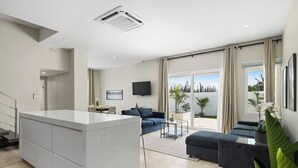 Ein modernes Wohnzimmer mit einer zentralen Kücheninsel, einer Sofagarnitur, einem Flachbildfernseher und einem großen Fenster mit Vorhängen.