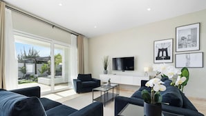 Ein modernes Wohnzimmer mit Sofa, Couchtisch und Fernseher. Ein Balkon mit Pflanzen bietet Blick auf die Stadt.