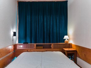 A bedroom with a wooden headboard, a large bed, a nightstand with a lamp, and a window with a blue curtain. - 7th floor, balcony, tv, 40m², Les Arcs (Les Arcs)