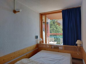 A bedroom with a bed, a window with a view of the outside, a wooden headboard, and a small table with a lamp.