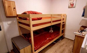 A bunk bed with a red mattress, a wooden bench, a small cabinet, and a wall-mounted picture.