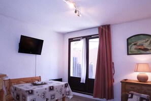 Una habitación con un televisor en la pared, una mesa con mantel estampado, una ventana con cortinas y una lámpara sobre una mesita de madera.
